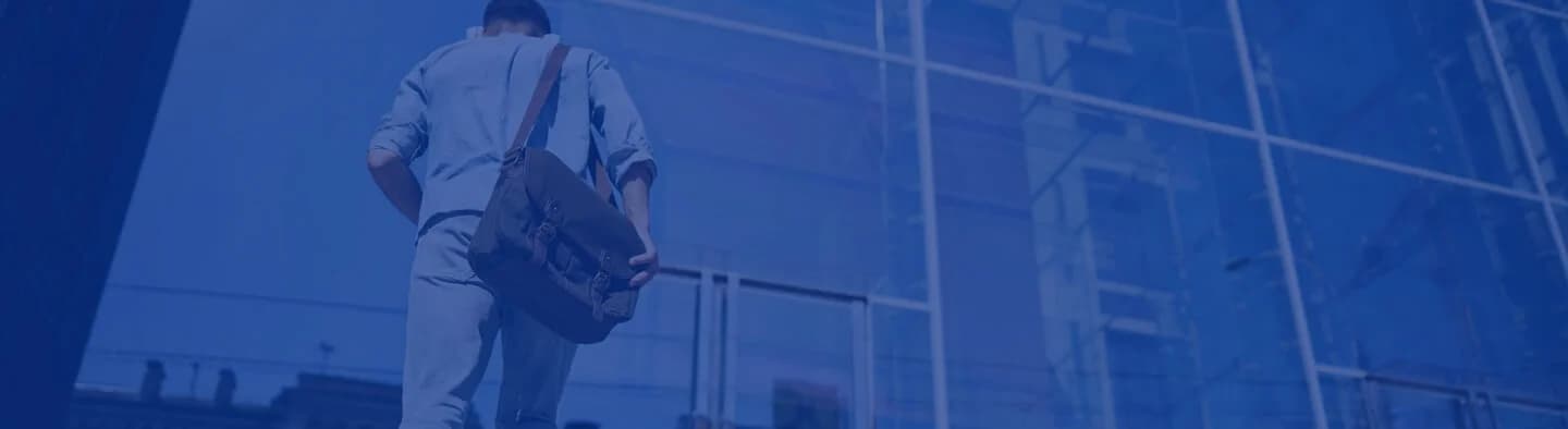 A man walking towards a glass building while carrying a bag.